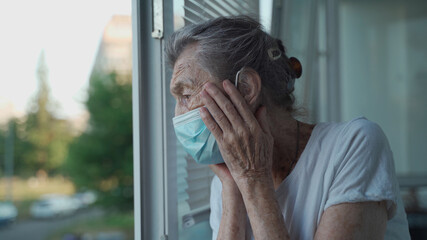Worried senior woman in a protective medical mask sadly looks out the window with her head clasped...