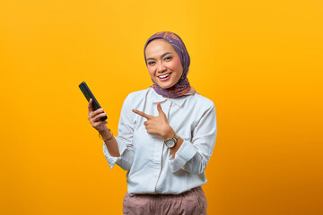 Portrait of smiling Asian woman pointing smartphone over yellow background