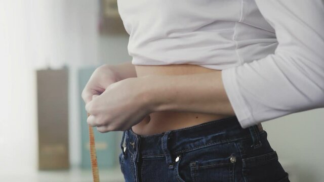 A young woman stands in front of a mirror and measures the circumference of her waist with a tape measure. Close-up