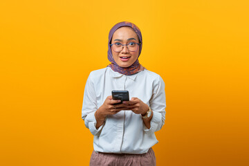 Shocked Asian woman using mobile phone and looking at camera over yellow background