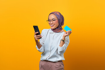 Cheerful Asian woman holding smartphone and showing blank card over yellow background