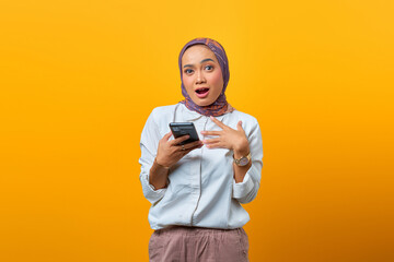 Portrait of surprised Asian woman using mobile phone with open mouth over yellow background