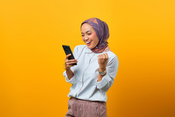 Excited Asian woman holding mobile phone celebrating luck over yellow background