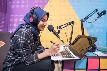 muslim woman is recording a podcast in studio with laptop taking a note with pen