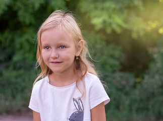 portrait of a child, a cute sweet girl 6-7 years old, blonde, looks ahead with a smile, against the background of a green park with bright rays of the sun