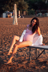 Obraz premium pregnant woman on the beach, full-length portrait of a brunette pregnant woman in a pink swimsuit posing on a bench on a sandy beach in summer at sunset. 