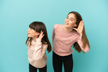 Little sisters girls isolated on blue background listening to something by putting hand on the ear