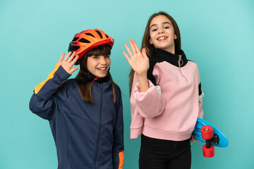 Little sisters practicing cycling and skater isolated on blue background saluting with hand with happy expression