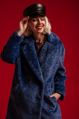 Happy smiling fashionable woman wearing trendy blue faux fur coat, leather cap, posing in studio,...