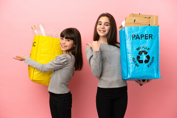 Little sisters recycling paper and plastic isolated on pink background extending hands to the side for inviting to come