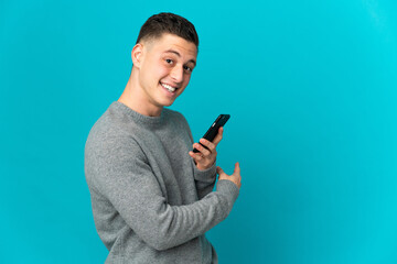 Young caucasian man isolated on blue background using mobile phone and pointing back