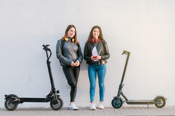 Two multiethnic female friends on electric scooters using smartphone outdoors. Two women standing over a white wall with headphones. Urban lifestyle and ecological transport concepts