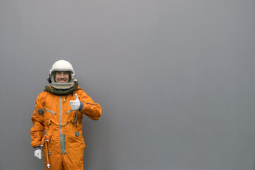 Happy astronaut wearing orange space suit and helmet showing thumbs up gesture on a gray wall background © Dmitry Zimin