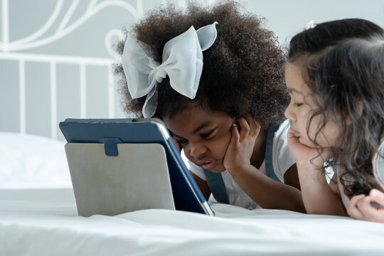 Selective focus on little African kid girl with bow tie on afro hair is lying on white bed put hands on chin and using tablet computer with Caucasian cute friend enjoy together at home
