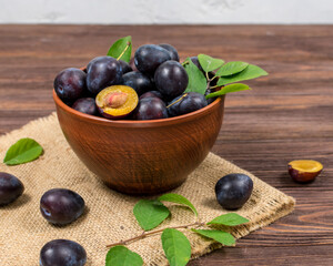 Fresh ripe plums whole and sliced with leaves in a bowl on burlap mats on a brown wooden table. Summer harvest.