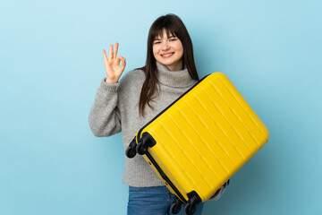 Young Ukrainian woman isolated on blue background in vacation with travel suitcase and making OK sign