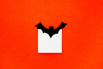 Crafts for the celebration of the concept of Halloween. Bat, cut out of paper and a white square, on an orange background. Copy space, place for text. Flat lay top view. Minimal Halloween background