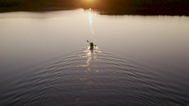 Following and approaching aerial FPV video of girl with red long hairs kayaking towards beautiful sunset, scenic Northern nature. Dark blue still water. Cinematic shot, sport vacation concept