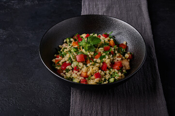 Close up lebanese or syrian traditional salad tabbouleh made of bulgur or couscous, poultry meat, parsley, mint in dark bowl with napkin on black background. Top view, copy space