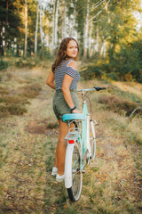 Fototapeta na wymiar Portrait of a young girl with a blue bicycle. Girl in the woods on a bicycle. Striped T-shirt and dark green shorts. Cycling in the woods