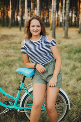 Obraz na płótnie Canvas Portrait of a young girl with a blue bicycle. Girl in the woods on a bicycle. Striped T-shirt and dark green shorts. Cycling in the woods