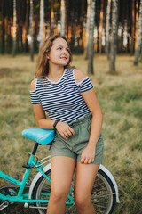 Obraz na płótnie Canvas Portrait of a young girl with a blue bicycle. Girl in the woods on a bicycle. Striped T-shirt and dark green shorts. Cycling in the woods