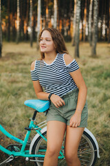 Obraz premium Portrait of a young girl with a blue bicycle. Girl in the woods on a bicycle. Striped T-shirt and dark green shorts. Cycling in the woods