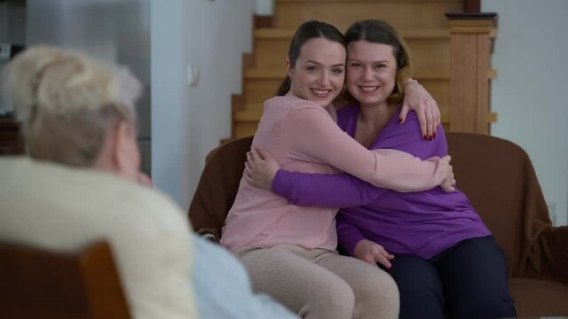 Happy mother and daughter hugging sitting on comfortable couch as unrecognizable grandmother talking. Joyful smiling Caucasian family at home indoors. Generations reunion concept