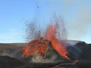 Volcanic vent at Fagradalsfjall, Iceland, erupting incandescent orange and red lava. Black cooled lava in the foreground and blue sky in the background.