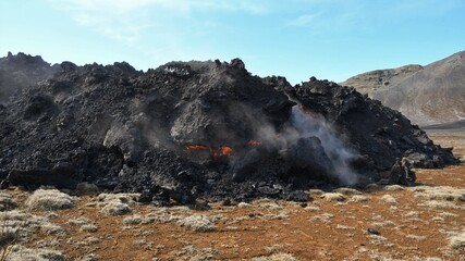 Lava flow at Fagradalsfjall, Iceland. Incandescent orange lava visible, within the black lava crust. Green and brown vegetation in the foreground. Sky and clouds in the background.