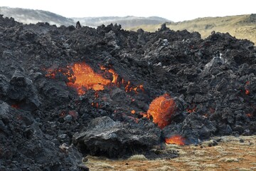 Lava flow at Fagradalsfjall, Iceland. Incandescent orange lava visible, within the black lava crust. Green and brown vegetation in the foreground. Sky and clouds in the background.