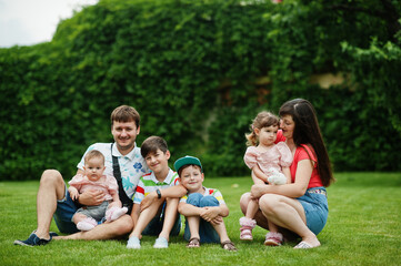Beautiful large family with young parents and four kids sitting in green grass at courtyard.