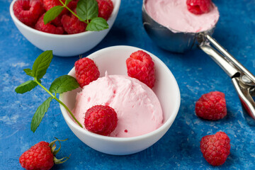 A scoop of homemade raspberry ice cream with fresh raspberries and mint in a white cup on a blue background. Delicious and romantic dessert. Close up