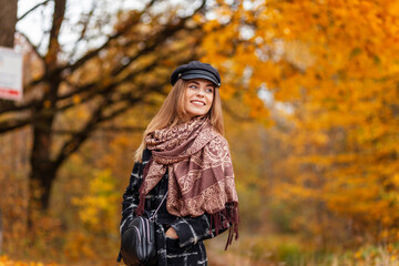 Plakat Autumn portrait of a beauty happy young girl with a hat and a scarf in a fashion coat with a handbag walks in the park with yellow fall foliage