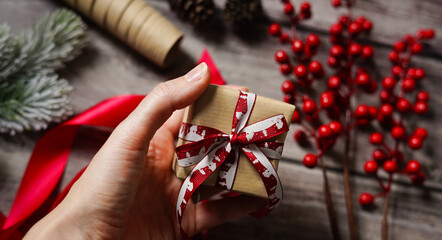 Woman's hand holding a present. Christmas composition with decoration, wrapping paper, red berries, ribbon and cones. Gift giving. Holiday craft gift wrapping. Merry Christmas.