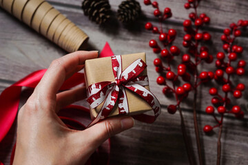 Woman's hand holding a present. Christmas composition with decoration, wrapping paper, red berries, ribbon and cones. Gift giving. Holiday craft gift wrapping. Merry Christmas.