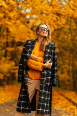 Happy stylish beautiful girl in fashionable vintage clothes with a knitted sweater and black coat with sunglasses walks outdoors with amazing yellow autumn leaves
