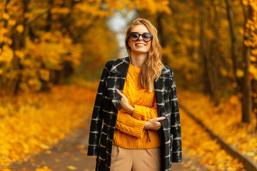 Happy beautiful european young woman with cute smile in fashion vintage clothes with knitted yellow sweater and coat wearing trendy sunglasses walks in autumn park with bright golden foliage.