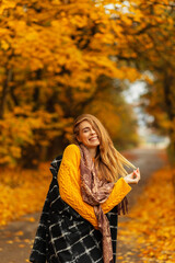 Happy Caucasian woman with beautiful smile in fashion clothes with black coat, knitted yellow sweater and scarf enjoy walks outdoors with amazing colorful autumn foliage. Funny girl emotions