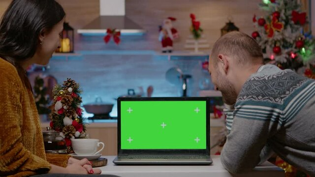 Festive couple looking at horizontal green screen on laptop for christmas eve celebration. People with chroma key and isolated mockup template preparing for seasonal holiday festivity.