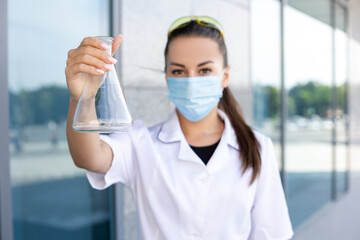 Scienсe, biology, chemistry and medicine concept - European smiling female biologist in medical gown and protective mask shows Erlenmeyer flask and looks into camera outside.