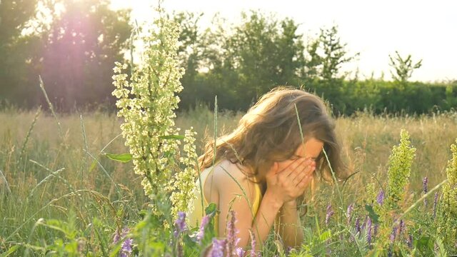 Pretty young girl is sitting in the middle of the field with flowers during summer time and sneezing, hypersensitivity, asthma, or allergy concept