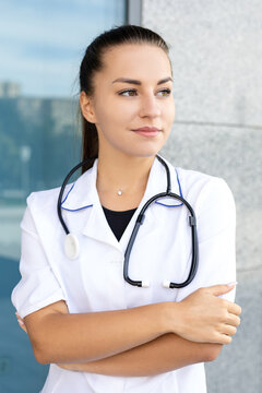 Medicine, healthcare, profession and people concept - portrait of a European white female doctor crossing her arms in a medical gown, wearing a stethoscope outside in sunny weather, looking away