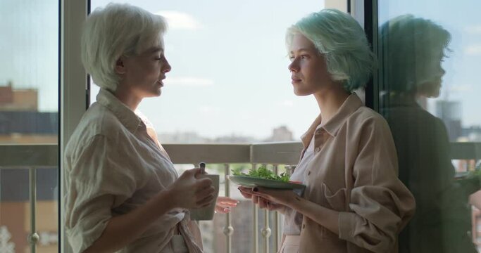 Two Cheerful Women Lesbians Holding Plate Of Breakfast And Talking In Balcony, Laughing, Having Nice Day Or Morning At Weekends, Lesbian Couple Spending Their Day Together. Side View, Copy Space