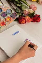 hand of a person holding a brush and painting with colors on a blank canvas, next to it is a palette and bouquet of colorful roses, objects to paint and rose buds, studio