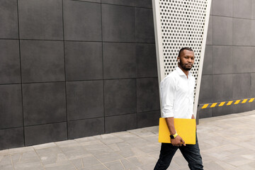 Young adult male student or freelancer in stylish clothes standing with yellow laptop outdoors, walking to the office for business meeting. Serious successful businessman looking at the camera, posing