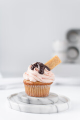 Vanilla cupcake with a tall swirl of pink frosting, chocolate glaze and a cookie, plain cupcake with pastel pink buttercream with chocolate and cookie garnish