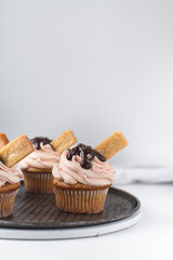 Vanilla cupcake with a tall swirl of pink frosting, chocolate glaze and a cookie, plain cupcake with pastel pink buttercream with chocolate and cookie garnish