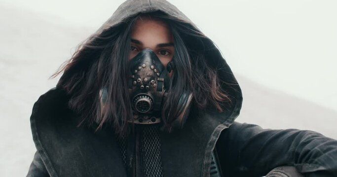 Close-up portrait post apocalyptic woman wearing grunge clothes hooded black mantle protective gas mask respiratory sitting on white hill in desert. Cyberpunk warrior, future female survivor