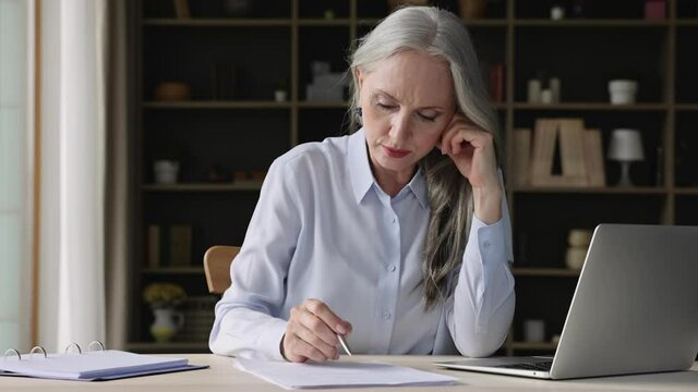 Focused 50s businesswoman sit at desk use laptop, looking through papers documents, attorney works with contract, check correspondence, report, analyzing data and agreement details, paperwork concept